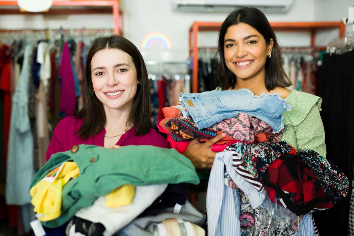 Two women in a thrift store, holding piles of of clothing