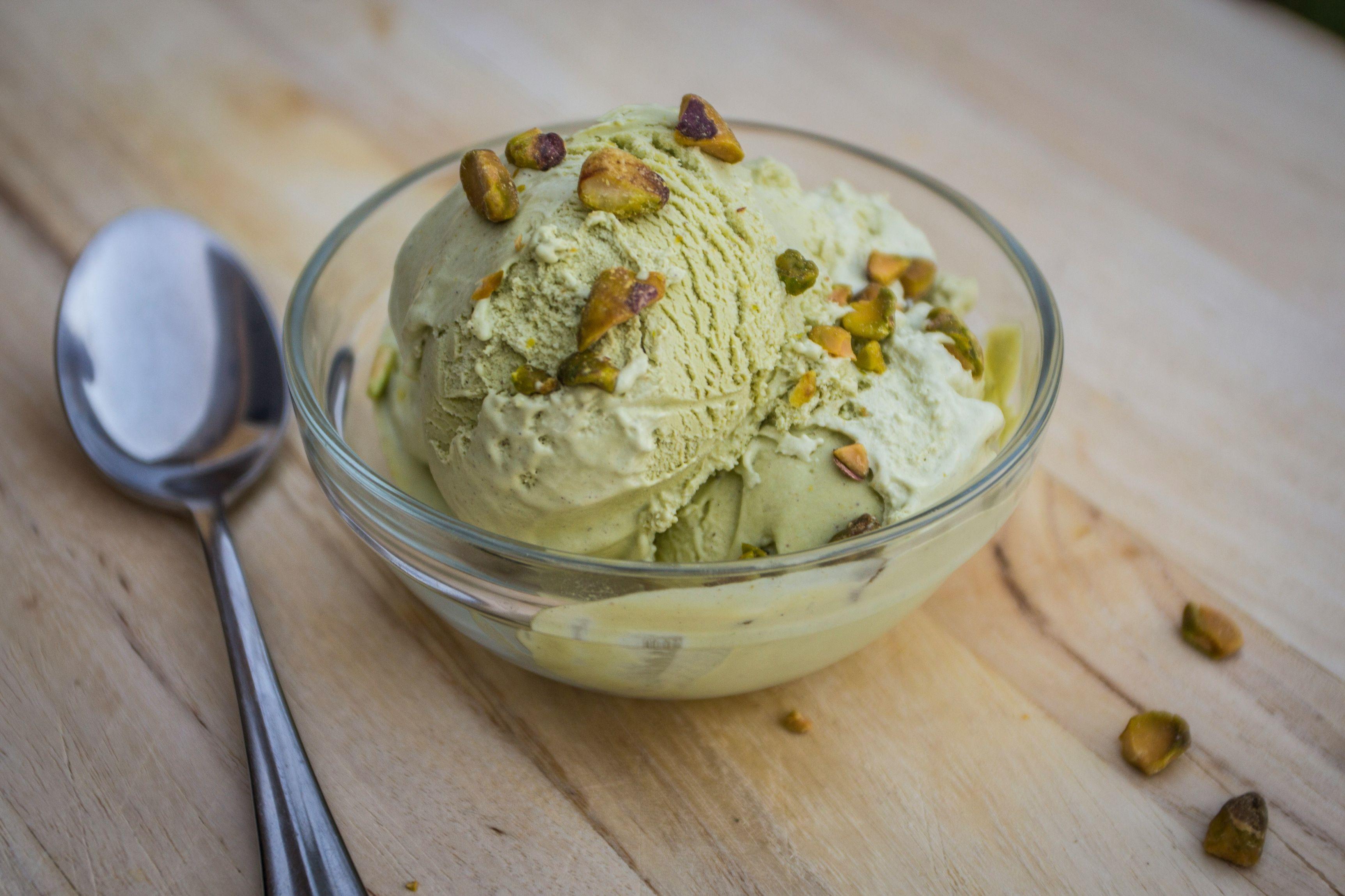 A bowl of pistachio ice cream is pictured topped with pistachios, with pistachio nuts and a spoon beside the bowl.