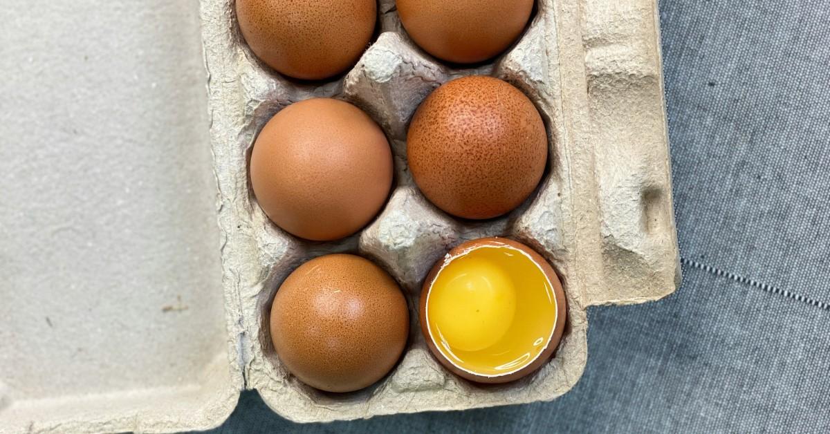 A carton of brown eggs with one egg cracked open showing the yolk