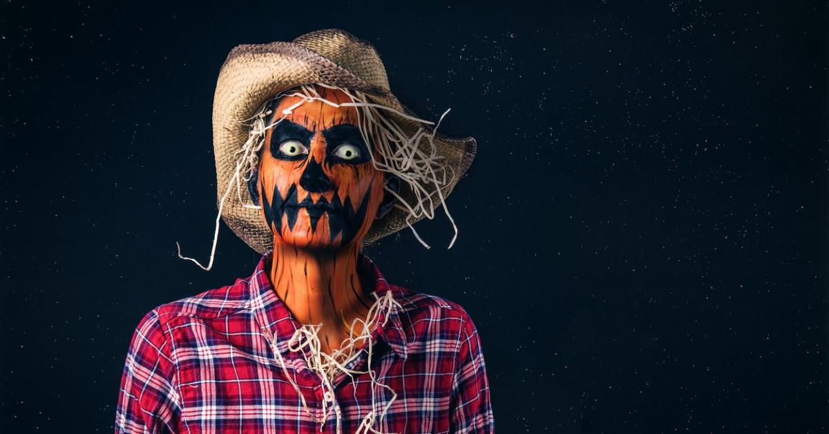A person wearing orange face paint dresses up as a spooky scarecrow