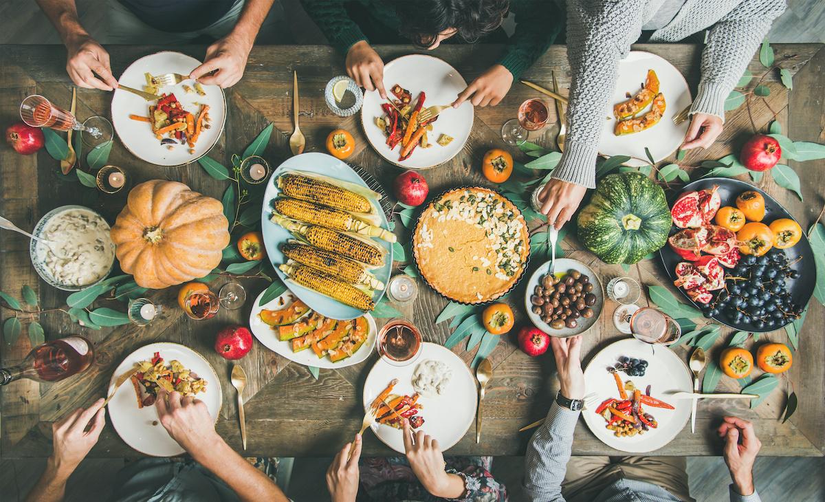 A Thanksgiving dinner on a wooden table featuring six people, pumpkins, corn, pumpkin pies, and more.