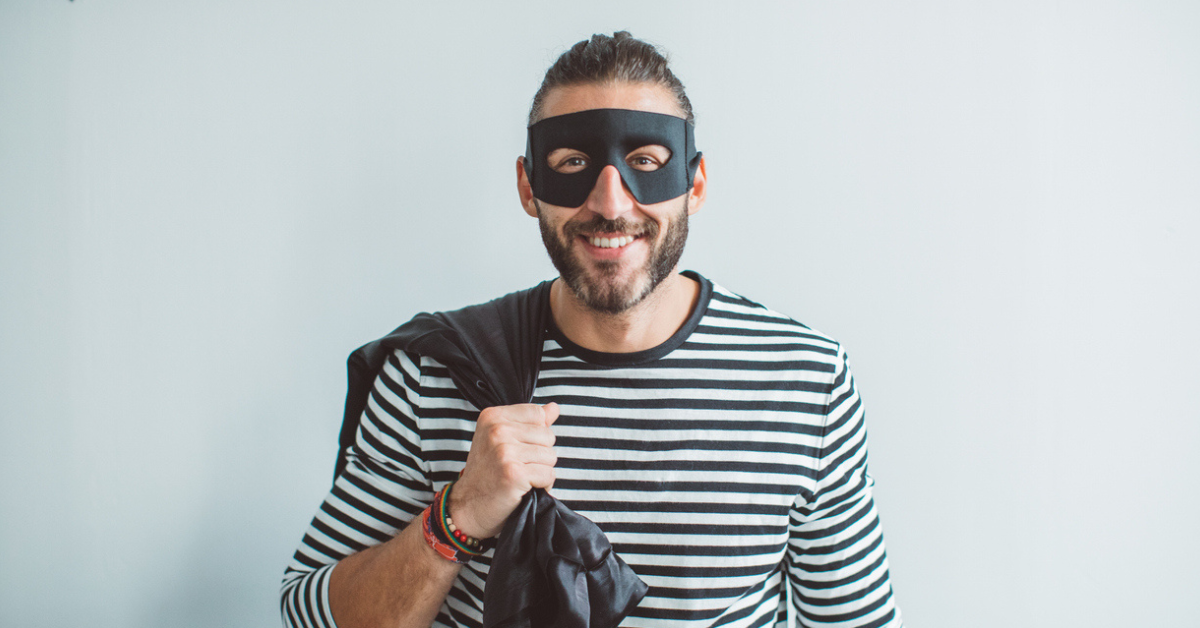 A man wearing a black bandit mask, black and white striped shirt, and carrying a black sack while dressed like a burglar