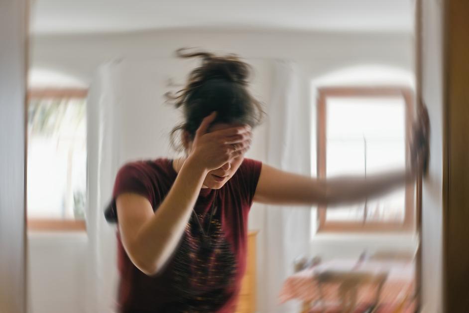 Blurry image of a woman in a red shirt holding onto the doorframe for stability as she holds her forehead in pain.